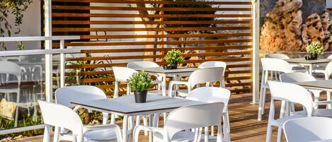 An outdoor seating area with white plastic chairs and tables, a wooden slatted partition, and a rock wall in the background.