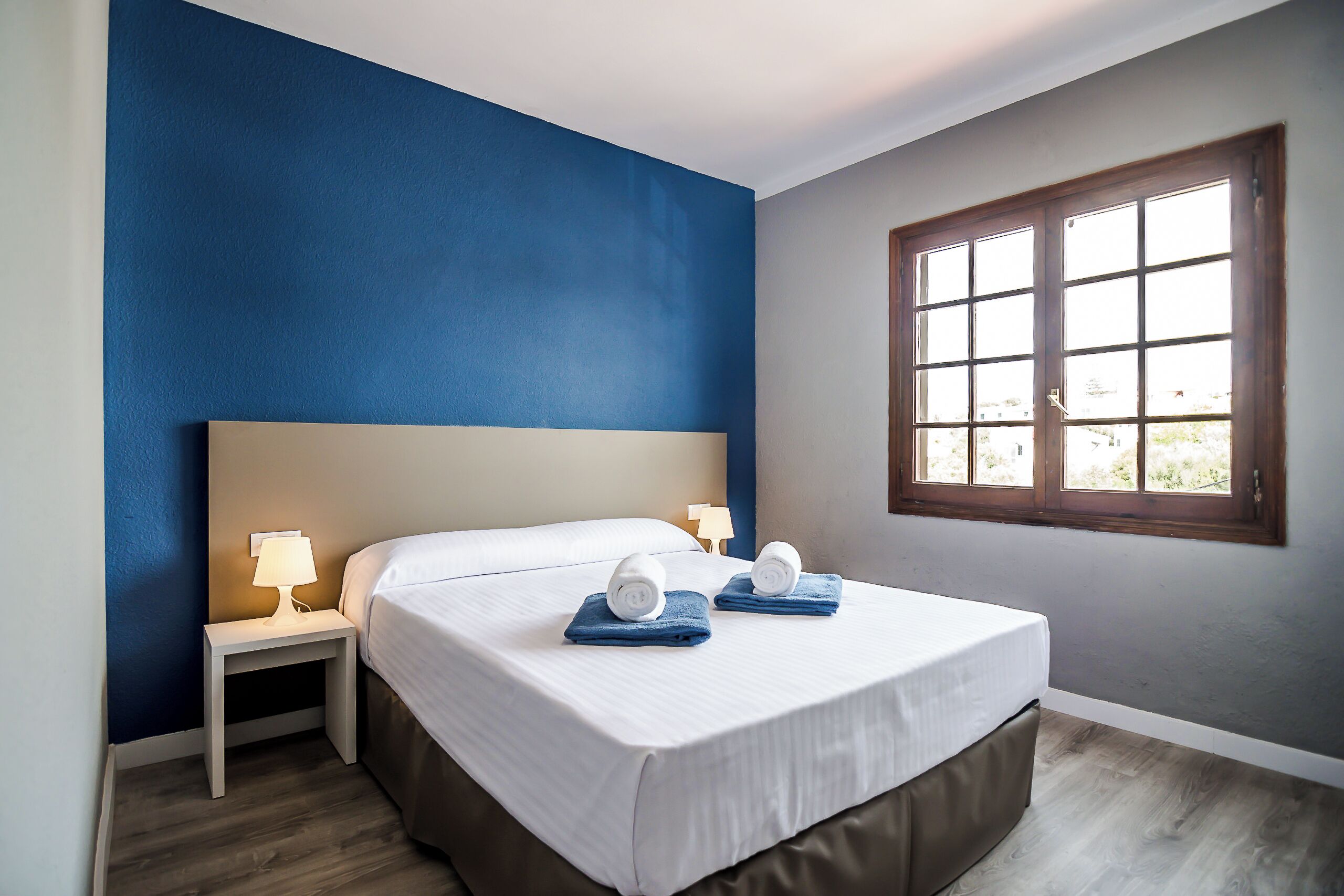 A bedroom with a large bed, two bedside tables with lamps, a window with a view, and a blue accent wall.