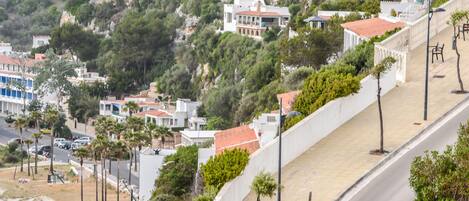 A coastal residential area with houses on a hillside, a paved walkway, and a road with parked cars.