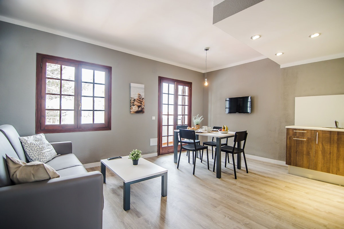 A modern living room with a sofa, dining table, and chairs. There is a kitchen area with cabinets and a TV.