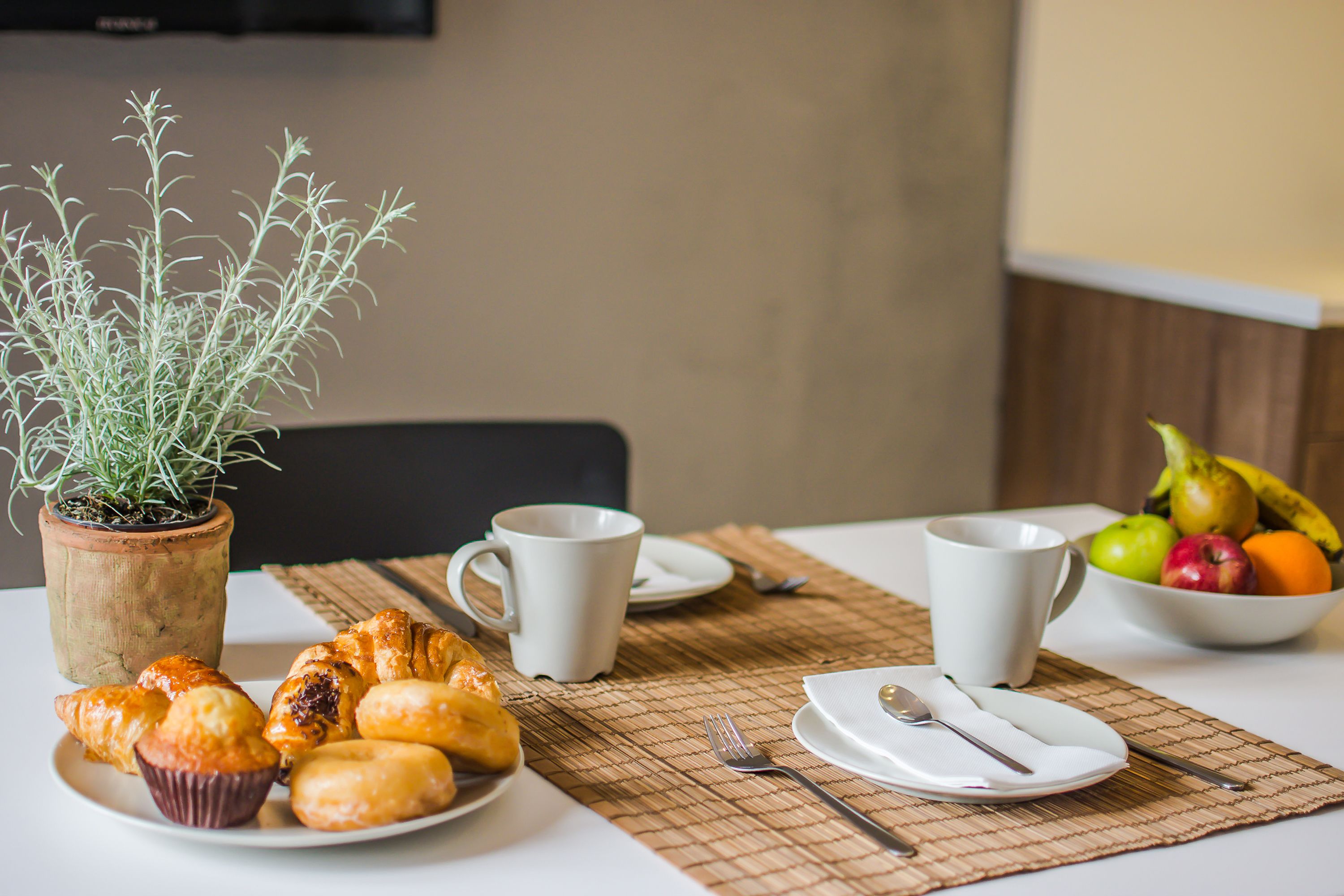 Un desayuno con pastelería, una taza de café, un bol de fruta y una planta en maceta.