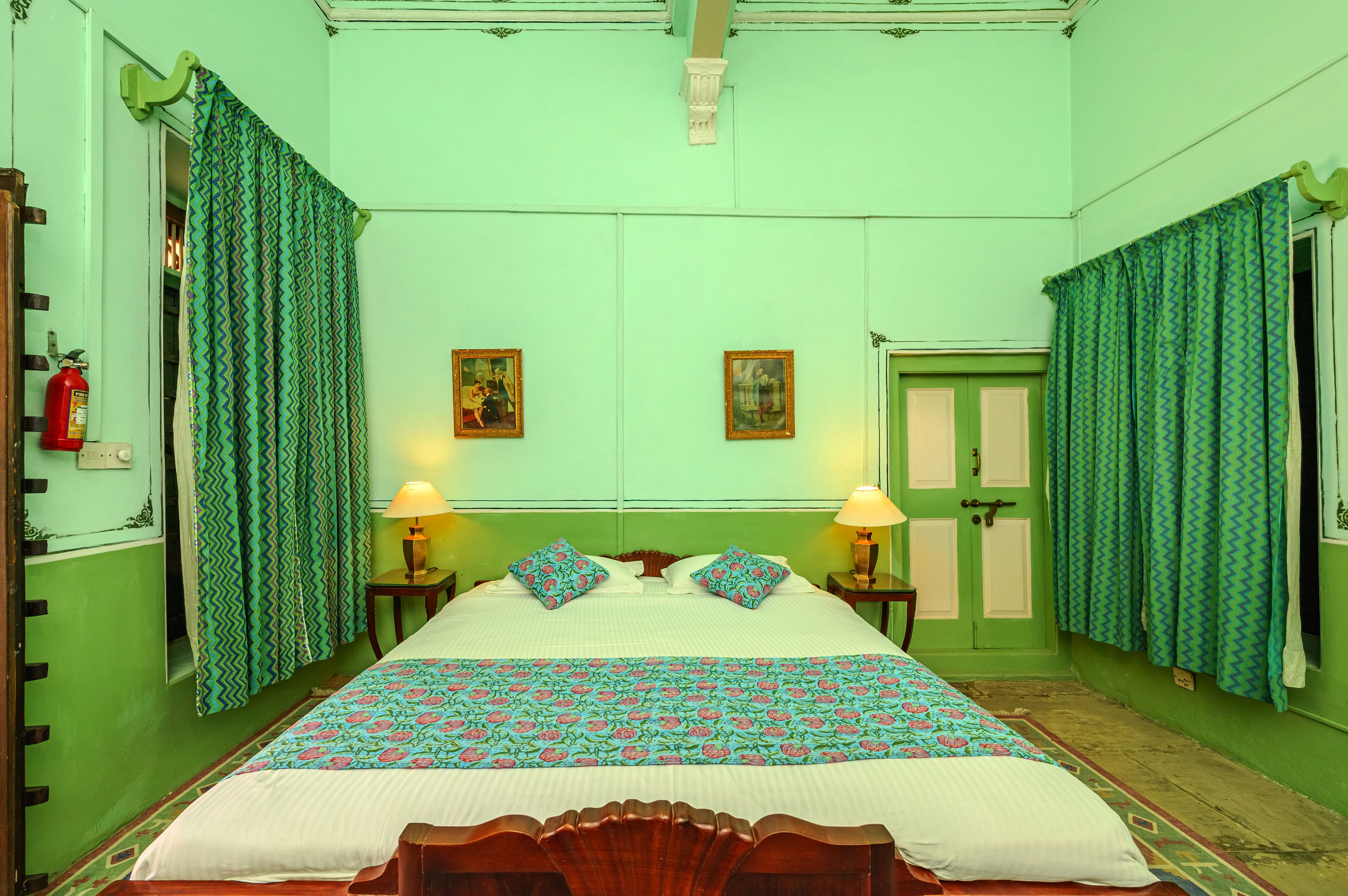 A room with a bed, two bedside lamps, a fire extinguisher, and two framed pictures on the wall.