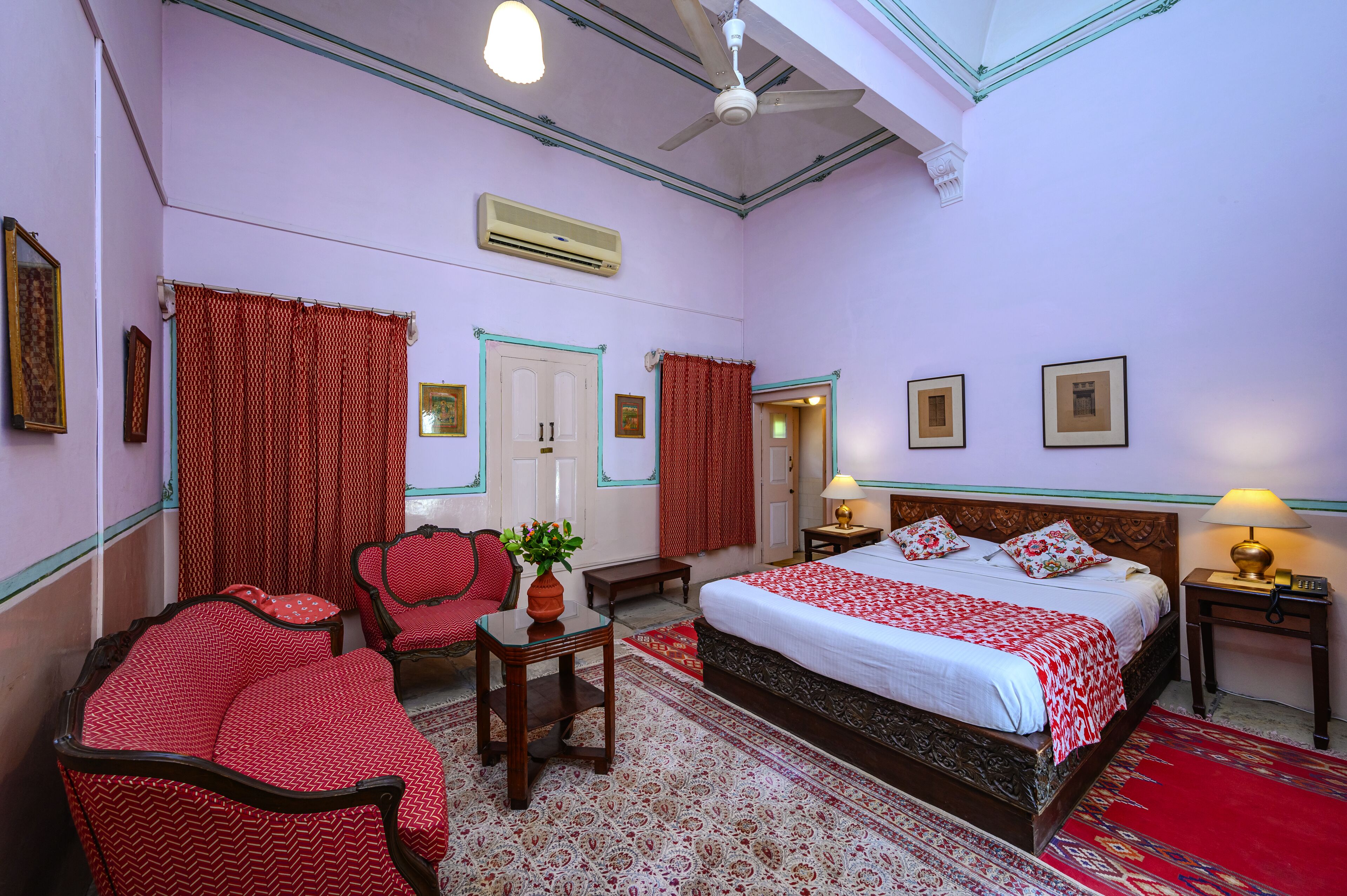 A hotel room with a large bed, two chairs, a small table with a plant, a red and white patterned rug, and a ceiling fan.