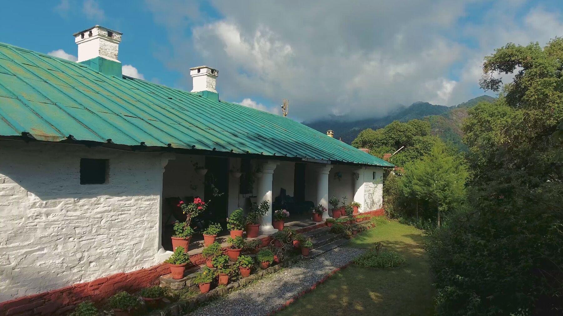 A white building with a green roof, surrounded by potted plants and a gravel path.