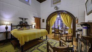 A traditional room with a bed, a desk, and a chair. There is a window with curtains, a fan, and a small table with a lamp. - Neemrana's - Ramgarh Bungalows (Kosiyakutoli)