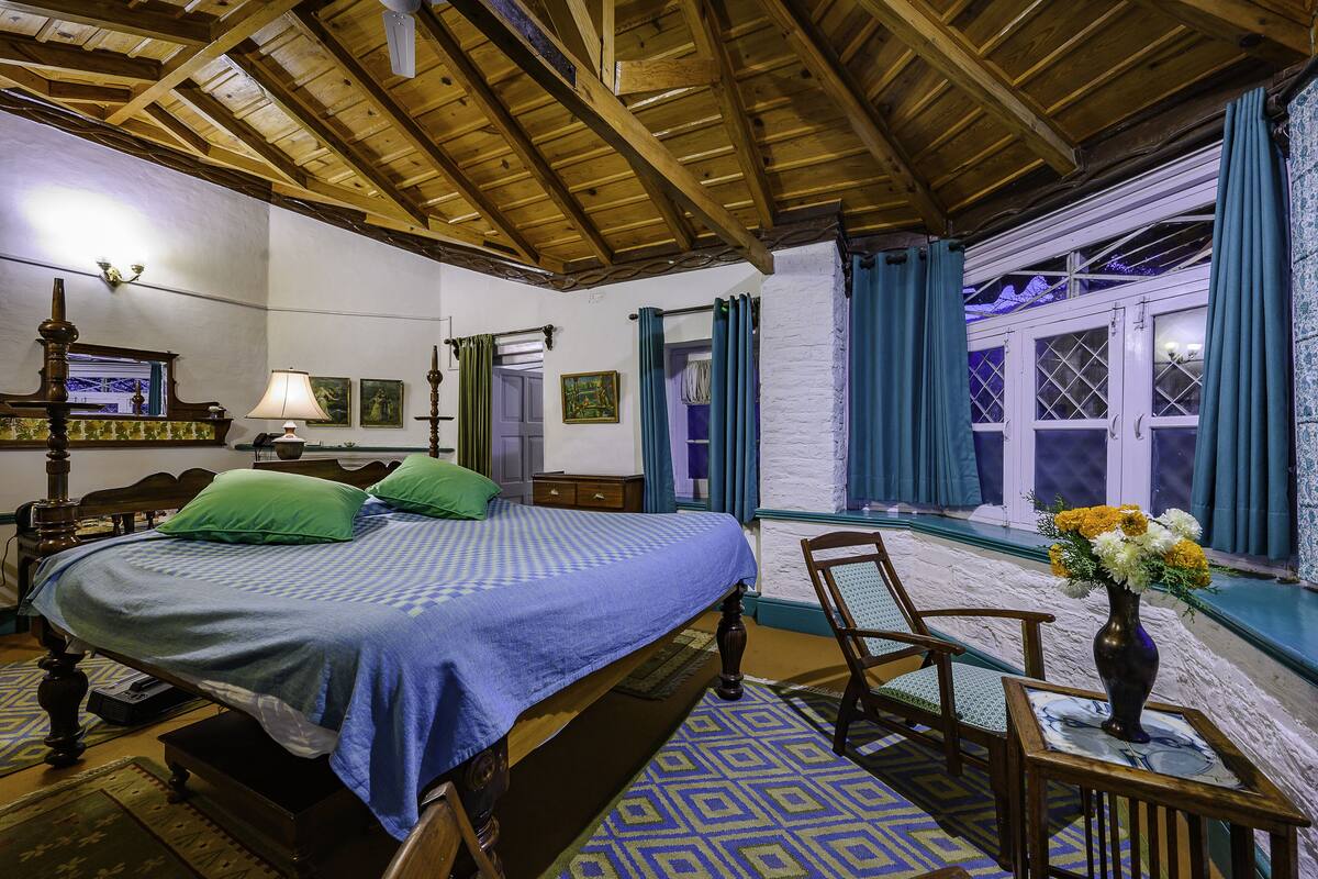 Una habitación con techo de madera, una cama con almohadas verdes, una silla, una mesa con un florero y una ventana con cortinas.