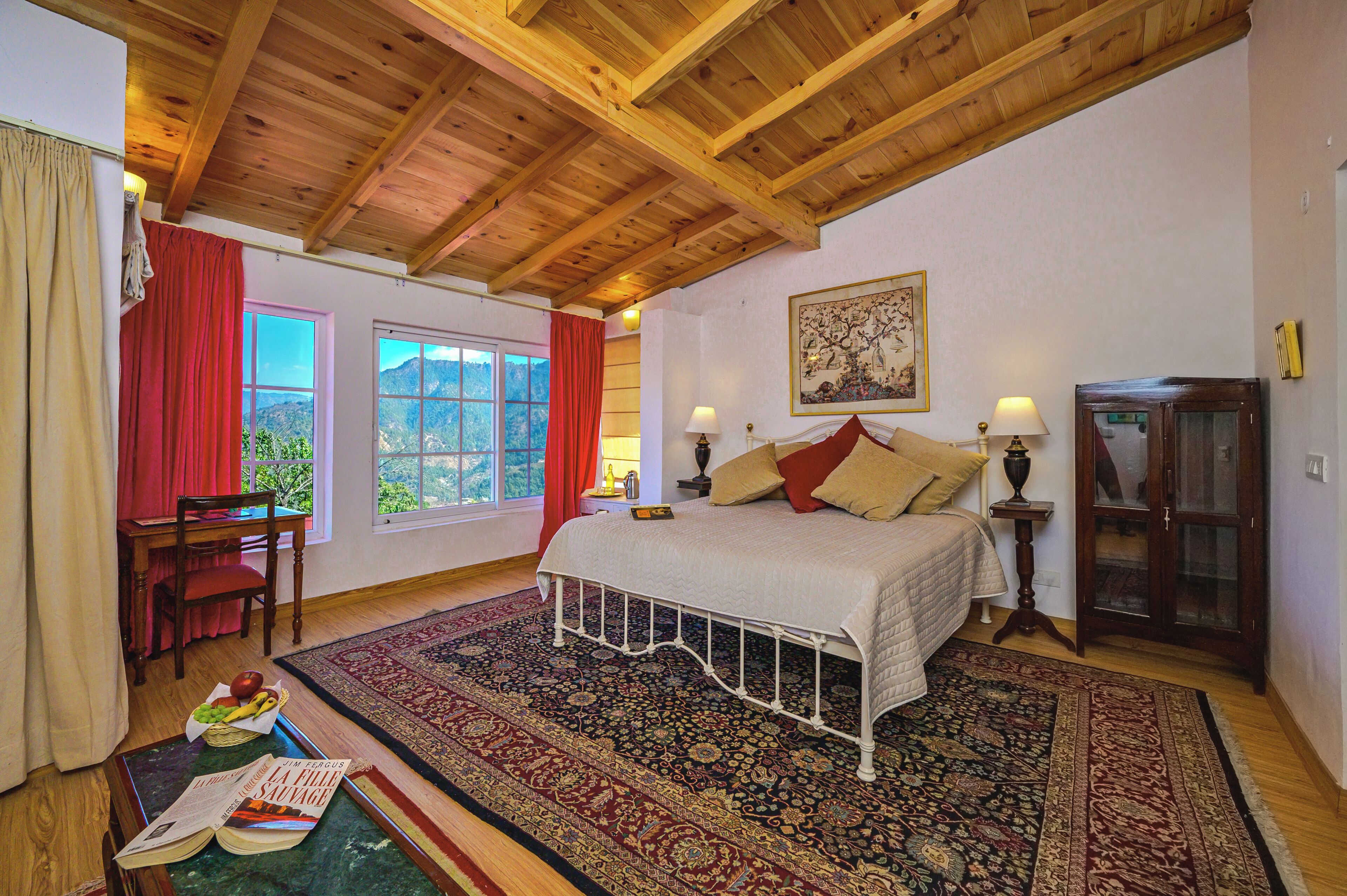 A bedroom with a wooden ceiling, a large bed, a desk with a chair, a window with a view, and a traditional rug.