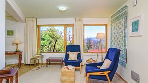 A room with two blue armchairs, a wooden side table, a small round table, and a bench. There are two windows with curtains, a lamp, and a wall-mounted air conditioner. - Neemrana's - Ramgarh Bungalows (Kosiyakutoli)