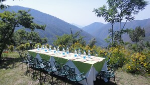 A table set outdoors with chairs, surrounded by mountains and greenery. - Neemrana's - Ramgarh Bungalows (Kosiyakutoli)