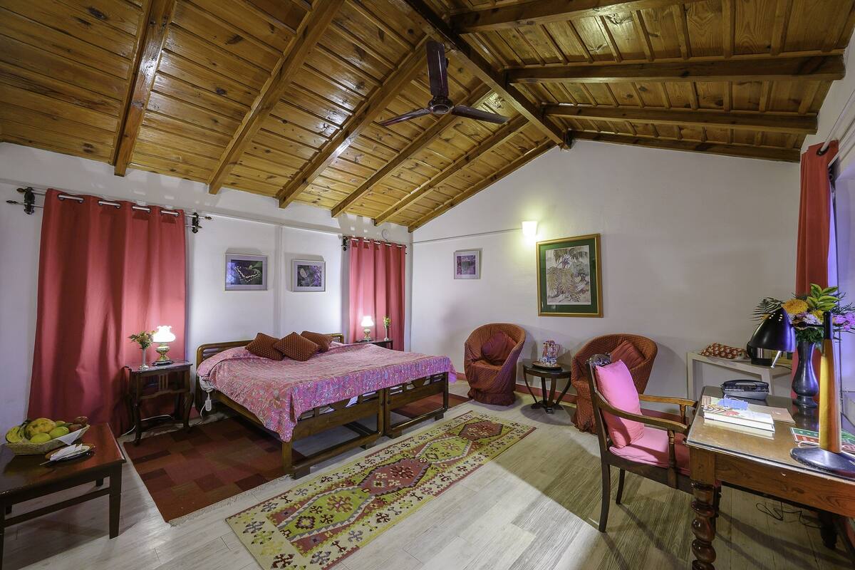 Un dormitorio acogedor con techo de madera, una cama con colcha rosa, una silla, un escritorio con lámpara y una alfombra.