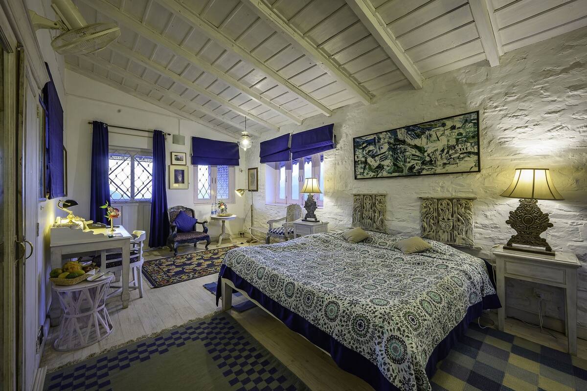 Un dormitorio con cama, escritorio, silla, lámpara y un cuadro en la pared.