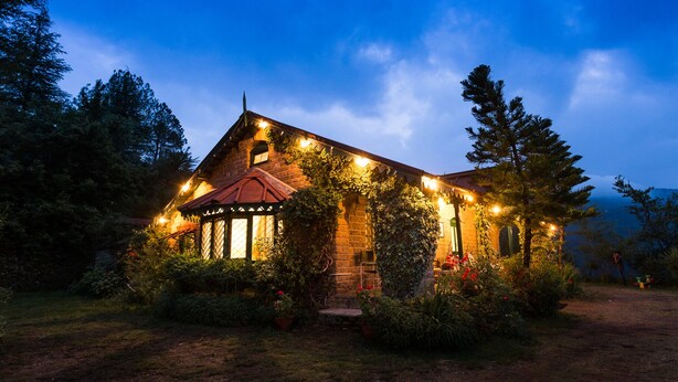 A rustic stone house with a red roof, surrounded by lush greenery and illuminated by string lights. - Neemrana's - Ramgarh Bungalows (Kosiyakutoli)
