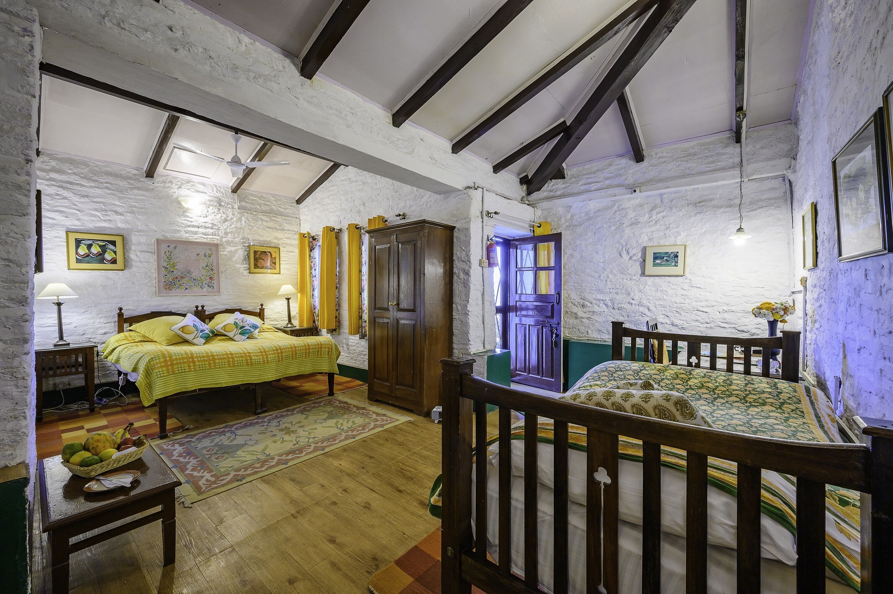Una habitación rústica con vigas de madera a la vista, una cama con cubrecama amarillo, un armario de madera y una mesita con una cesta de fruta.