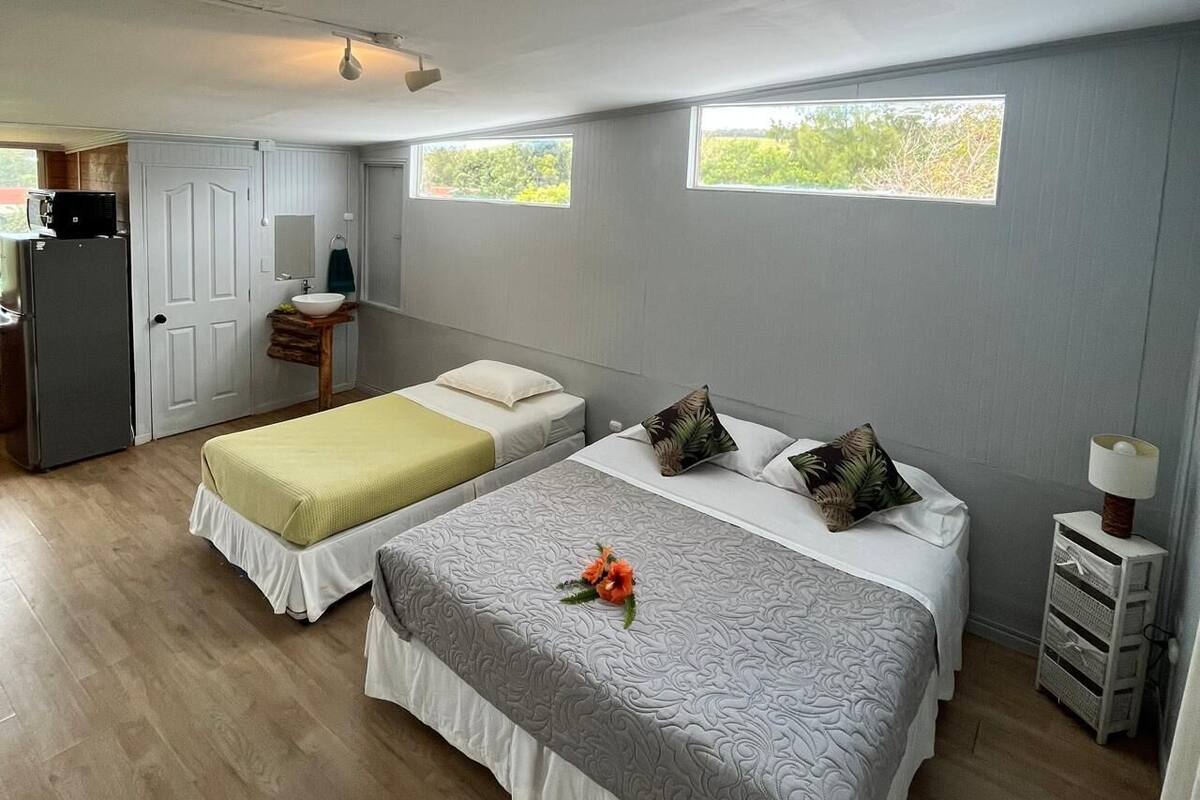 A bedroom with two beds, a small kitchenette, and a view of greenery through the windows.