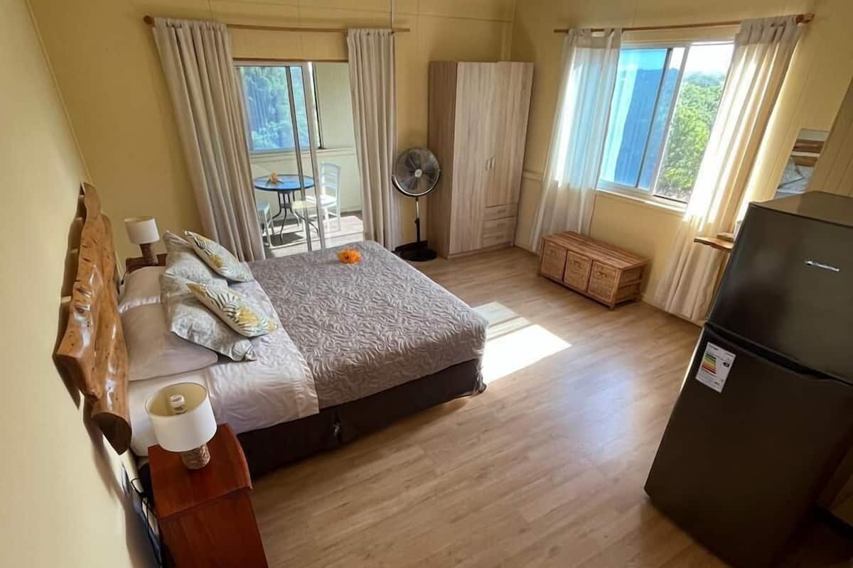 A bedroom with a bed, bedside table, lamp, and a fan. There is a window with curtains and a view of greenery outside.