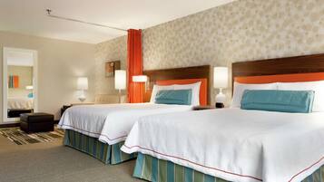 Une chambre d’hôtel avec deux lits, chacun doté d’une tête de lit en bois, de draps blancs et d’une jupe de lit à rayures vertes et bleues. On y trouve deux tables de chevet avec des lampes, un grand miroir et un papier peint à motifs.