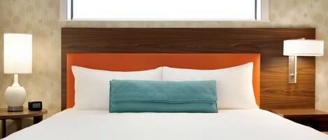 A hotel room with a large bed, a wooden headboard, a teal throw pillow, and a nightstand with a lamp.