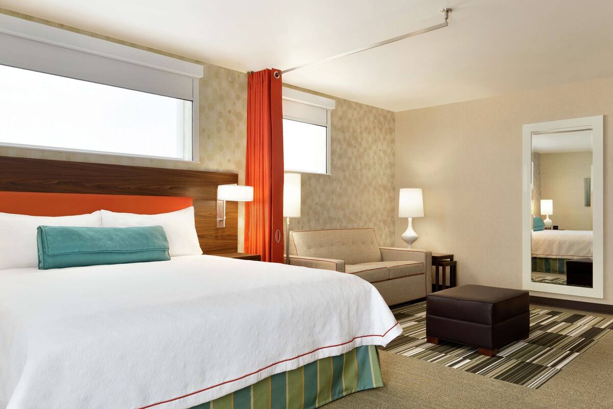 A hotel room with a large bed, a chair, a small table, a mirror, and a window with curtains.