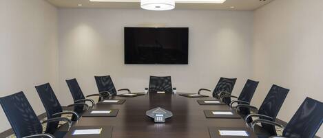 A conference room with a long table, black chairs, a central podium, and a large flat-screen TV on the wall.