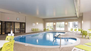 Una piscina interior con un reloj y una señal de salida en la pared, rodeada de sillas y mesas verdes.