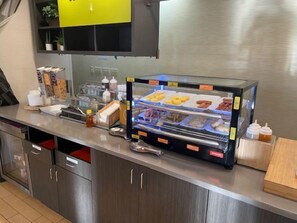 A bakery display case with various baked goods, a coffee machine, and condiments on a stainless steel counter. - Home2 Suites by Hilton Roanoke, VA (Roanoke)