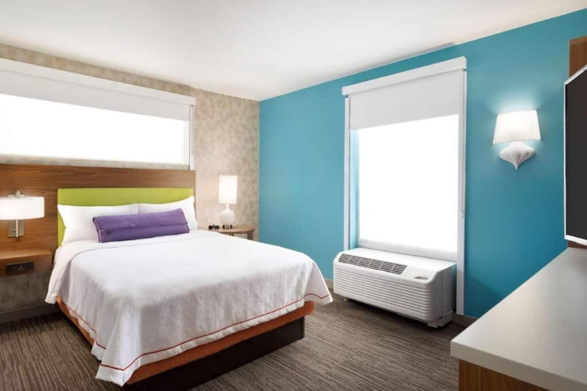 A hotel room with a bed, bedside tables, a lamp, a window, and an air conditioning unit.