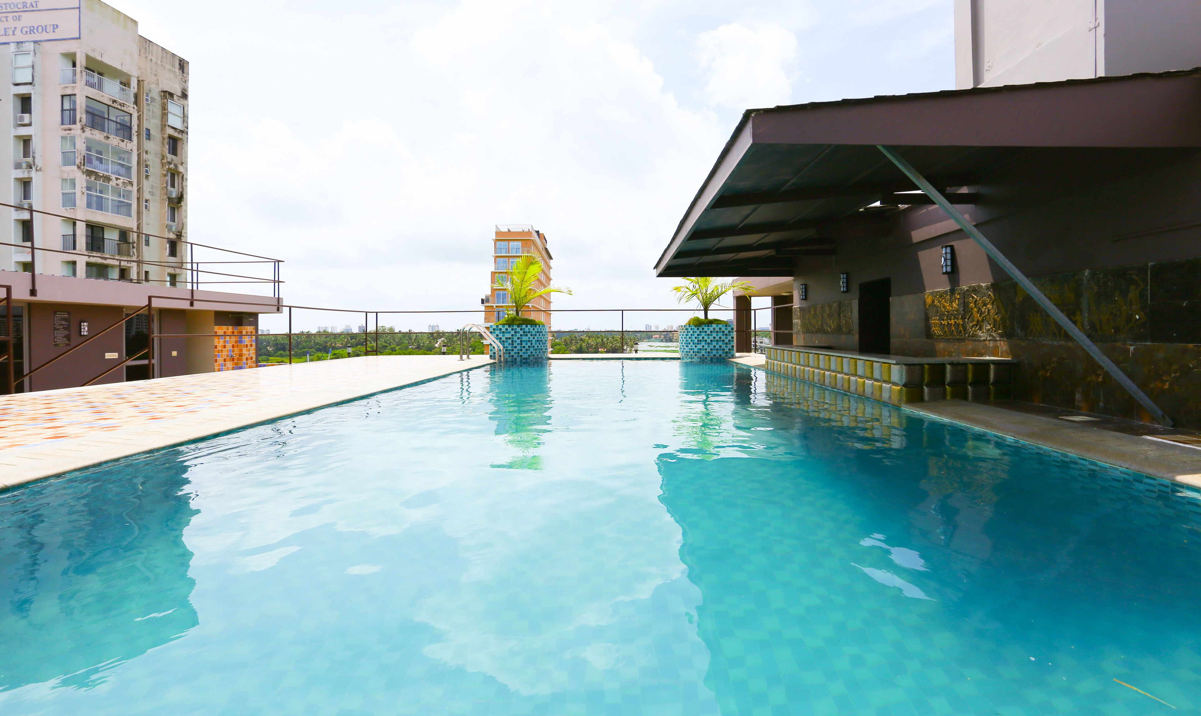 Una piscina con agua azul cristalina, rodeada por una solana con baldosas. Se ven dos palmeras y un edificio con balcón al fondo.