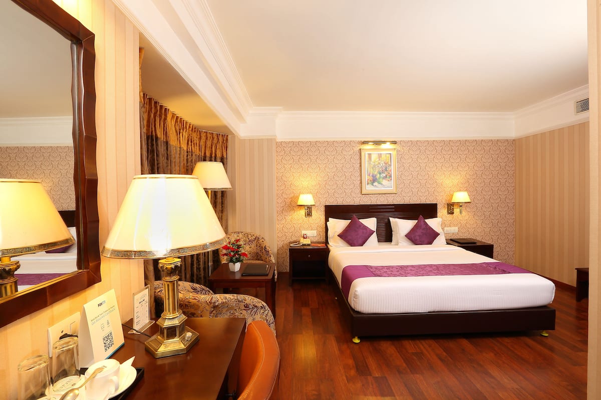 Quarto de hotel com uma cama grande, duas mesas de cabeceira, uma escrivaninha, uma cadeira, um espelho e uma luminária na parede.