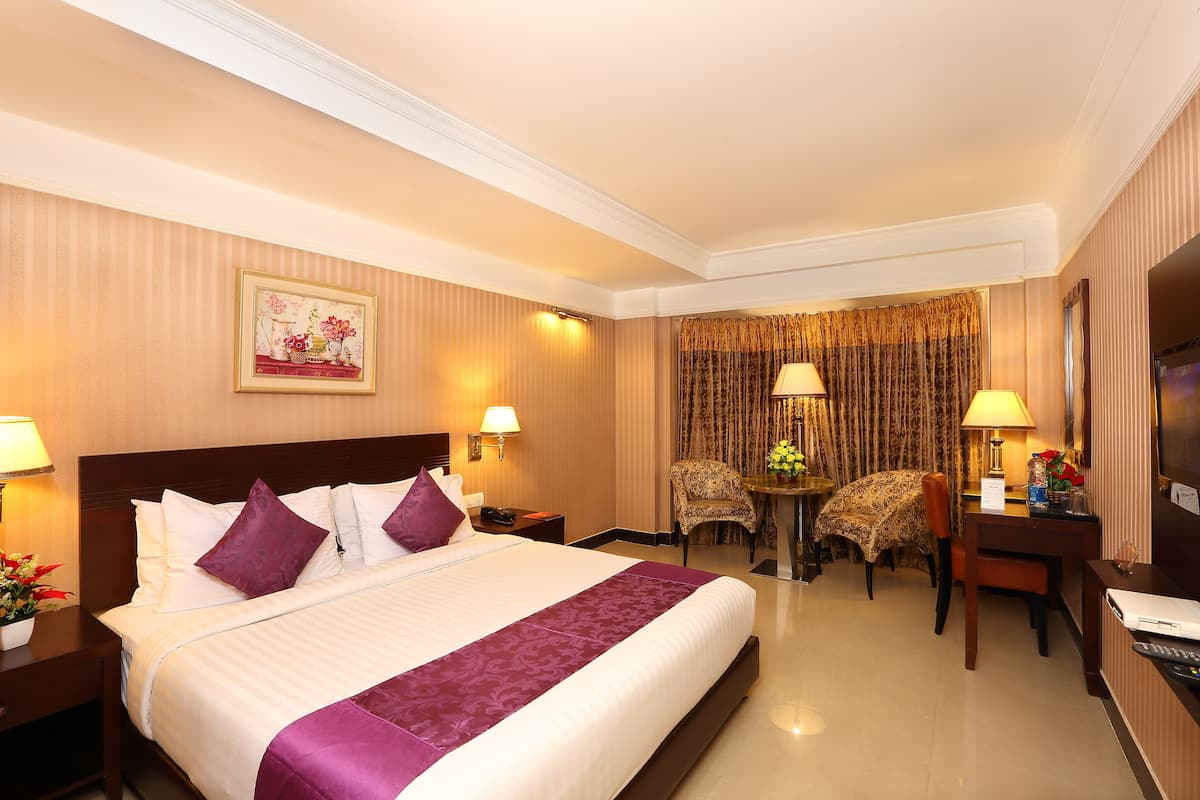 Quarto de hotel com uma cama grande, duas luminárias de cabeceira, uma mesa com cadeira, televisão e um quadro na parede.