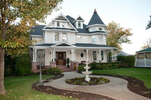 A large, two-story house with a wrap-around porch, a central fountain, and a brick foundation.