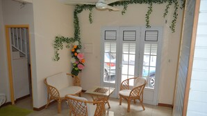 A room with a dining table, four wicker chairs, a potted plant with flowers, and a door with a view of parked cars outside. - Résidence Plocus (Fort-de-France)
