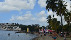A beach with palm trees, people swimming, and a large yellow inflatable structure. - Résidence Plocus (Fort-de-France)
