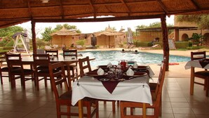 A dining area with a pool view, featuring tables set for dining, a thatched roof, and a pool with a person swimming. - Le Grand Calao (Ouagadougou)