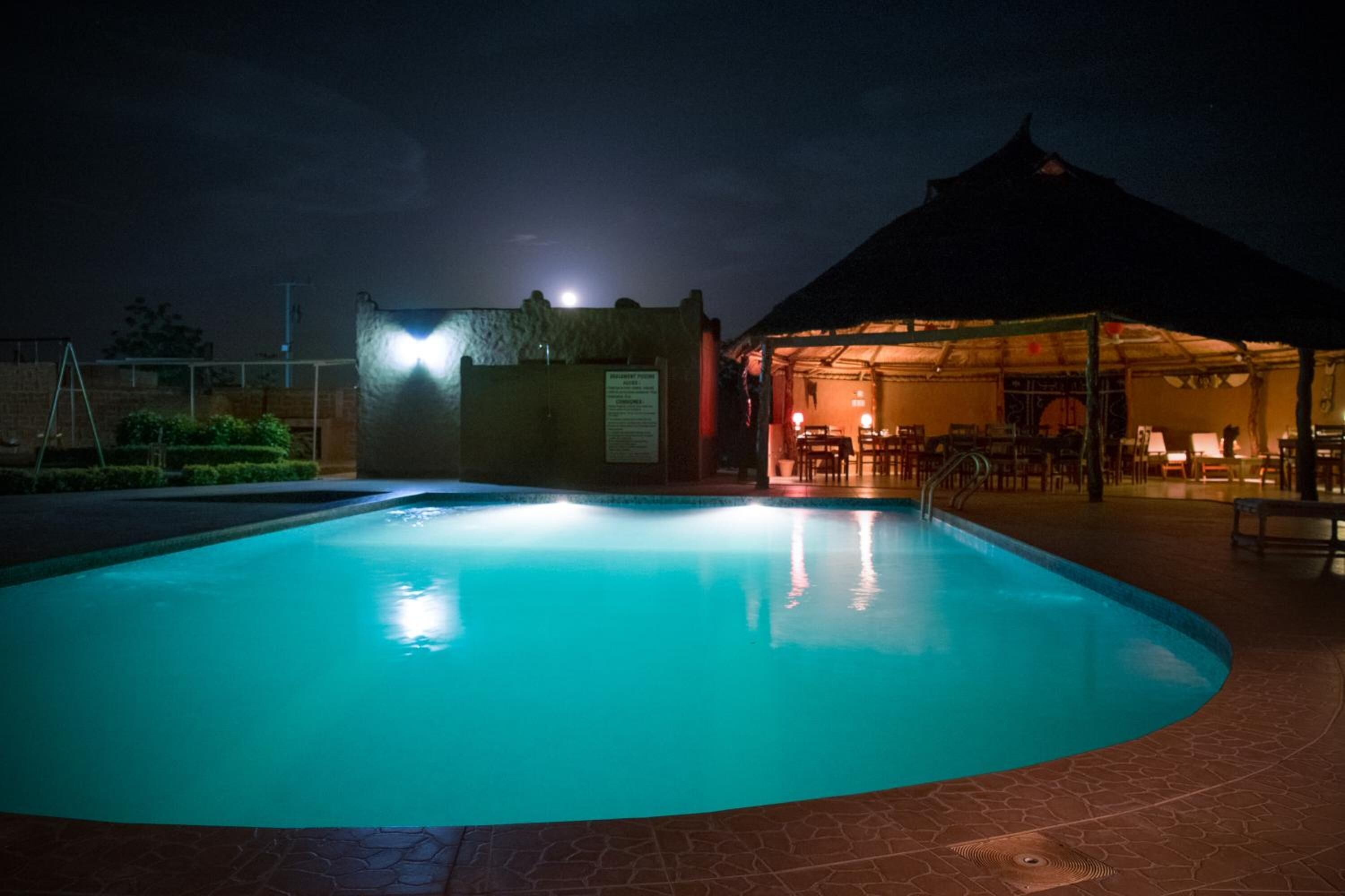 A swimming pool with a tiled deck, a covered seating area, and a building with a peaked roof in the background.