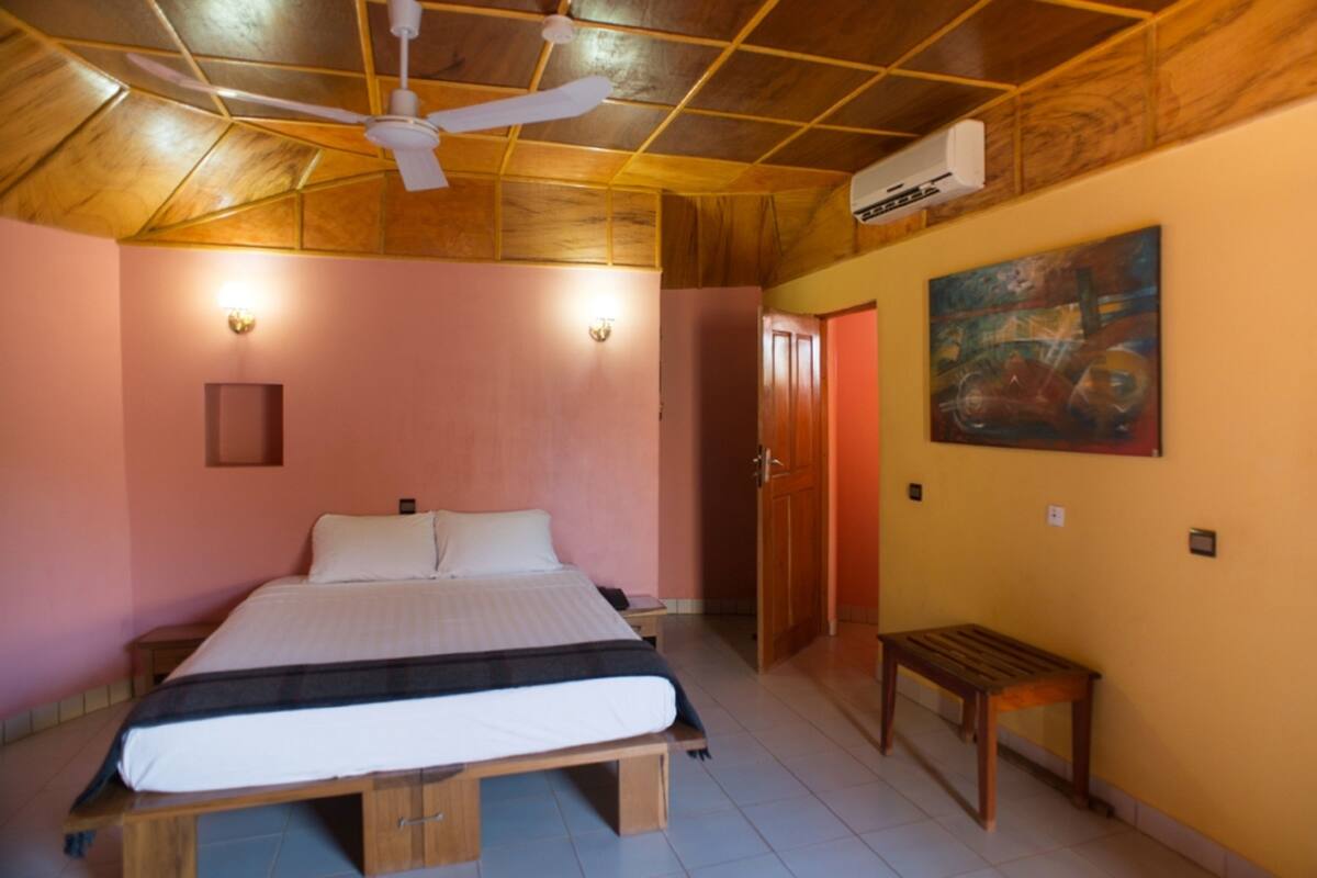 A bedroom with a wooden ceiling, a bed with a white mattress, a wooden bench, and a painting on the wall.