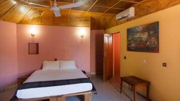 A bedroom with a wooden ceiling, a bed with a white mattress, a wooden bench, and a painting on the wall.
