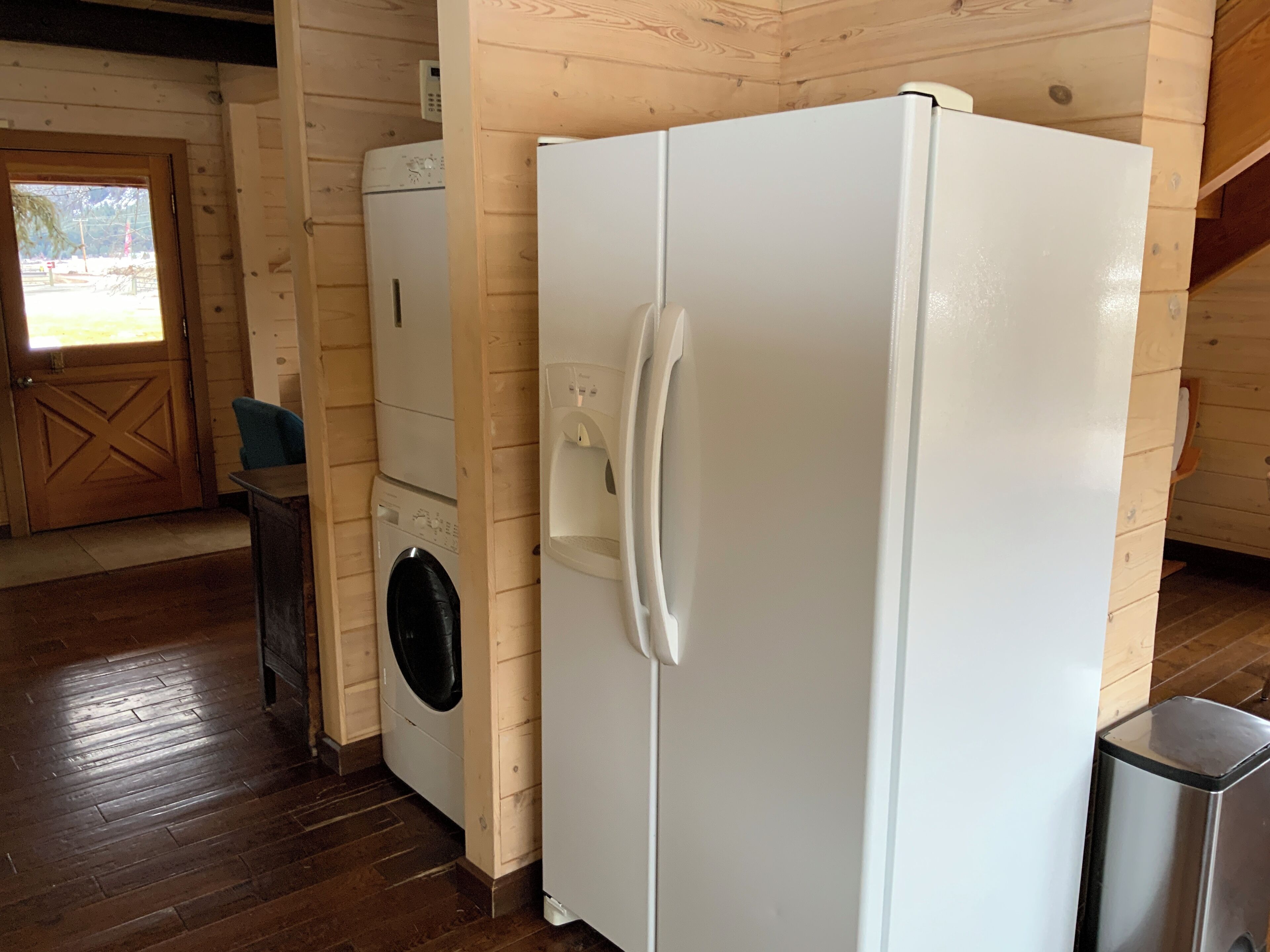 Une cuisine compacte aux murs et au sol en bois. Elle comprend un lave-linge, un réfrigérateur et une poubelle.