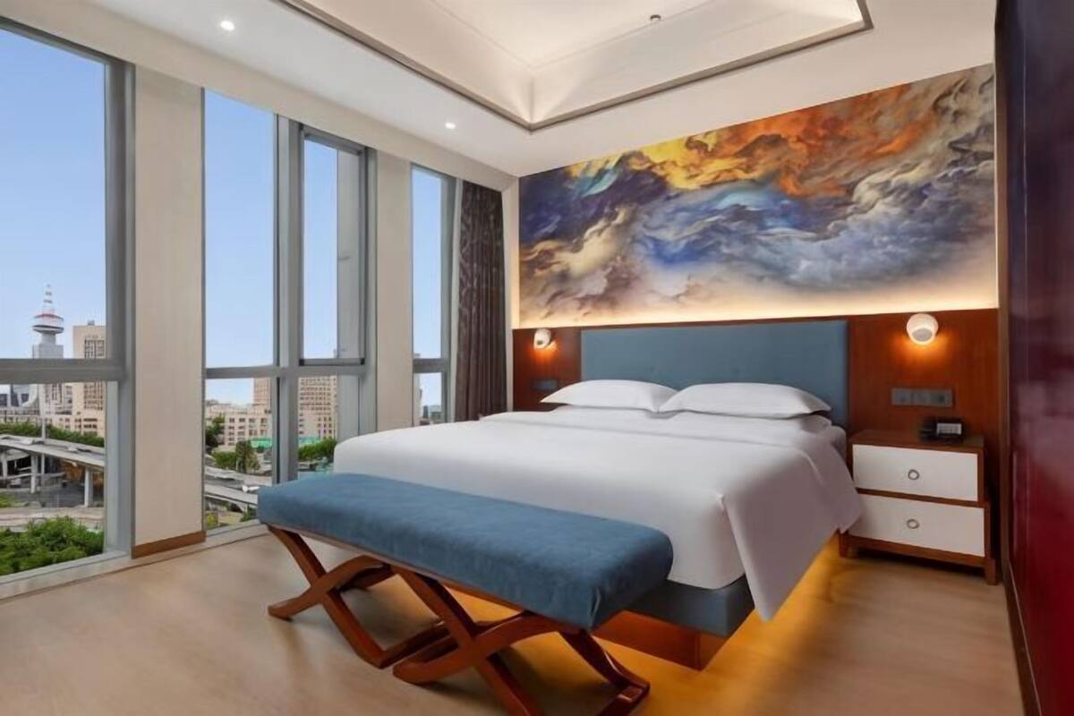 A modern hotel room with a large bed, a bench, a nightstand, and a city view through large windows.