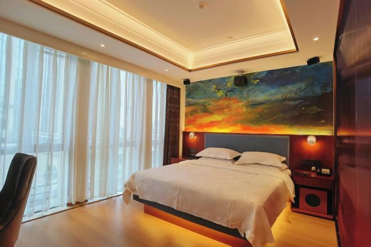 Habitación de hotel moderna con una cama grande, un cuadro sobre la cama, una silla y un amplio ventanal con cortinas.