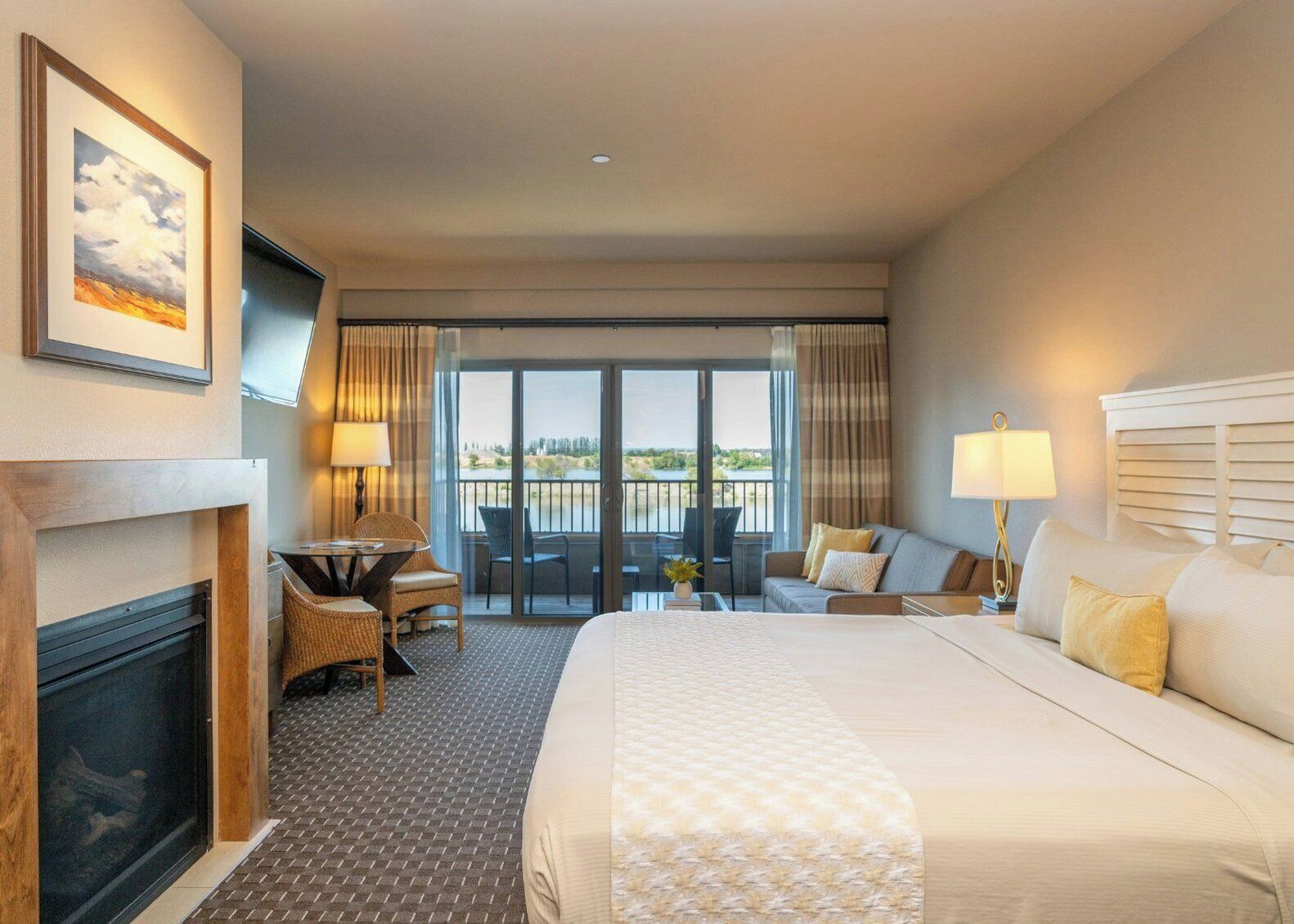 A hotel room with a large bed, a desk, a chair, a fireplace, and a balcony with a view.