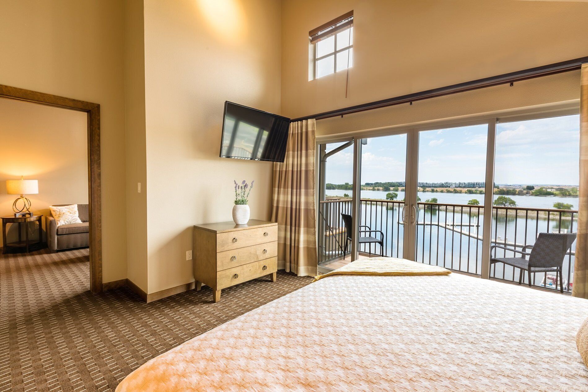 Habitación de hotel con una cama grande, un televisor montado en la pared, un aparador y vistas a un lago a través de amplios ventanales.