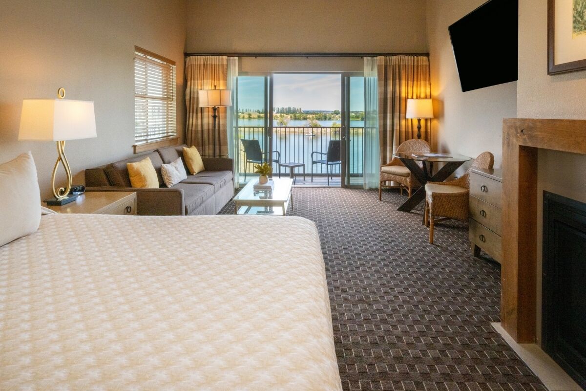 A hotel room with a large bed, a sofa, a small table, a fireplace, and a balcony with a view of a body of water.
