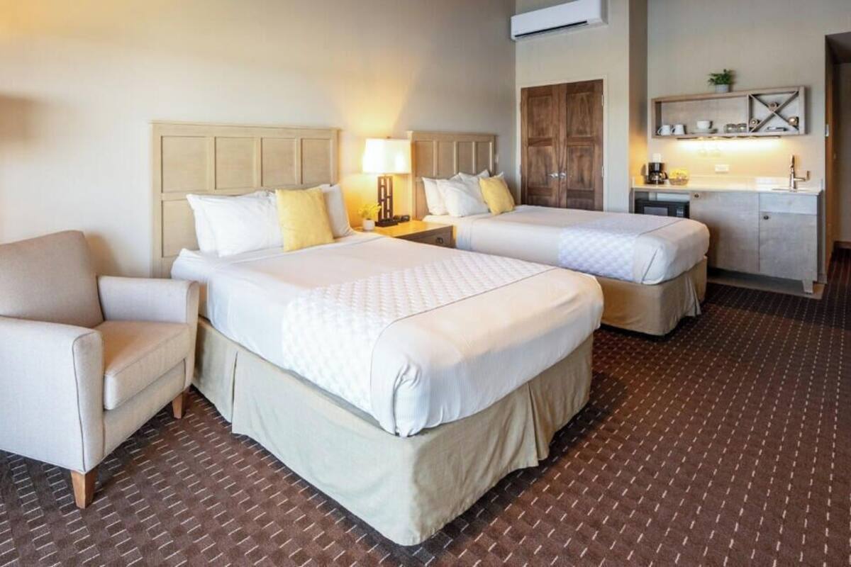 Habitación de hotel con dos camas, un escritorio, una silla, una lámpara y una kitchenette.