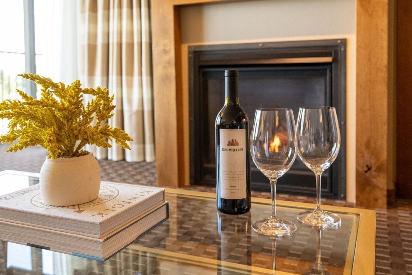 A book, a bottle of wine, and two glasses on a glass table with a fireplace in the background.