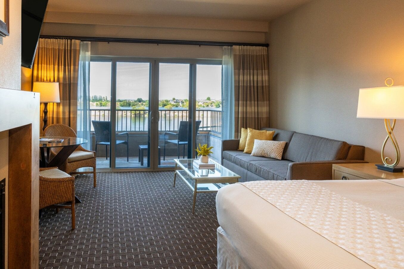 A hotel room with a bed, a sofa, a desk with a chair, a small table with a lamp, and a balcony with a view.
