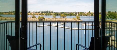 Un balcón con dos sillas y una pequeña mesa, con vistas a un lago tranquilo con un muelle y una orilla distante.