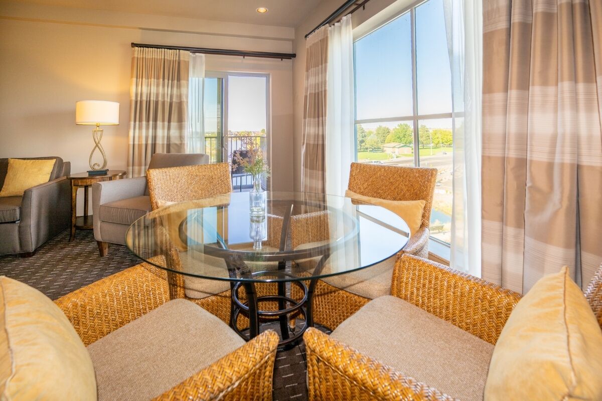 A modern hotel room with a glass-top dining table surrounded by wicker sofas, a grey sofa, and a small side table with a lamp. Large windows offer a view of the outdoors.