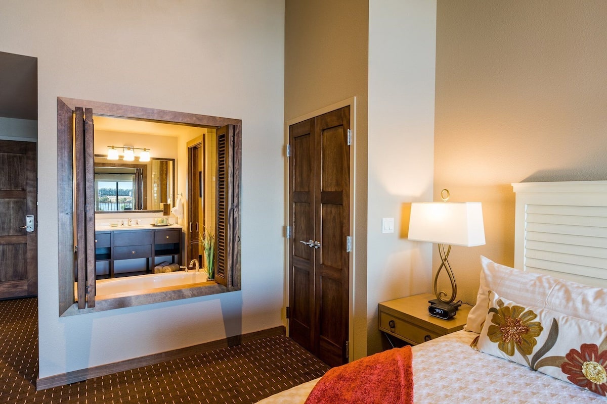 Habitación de hotel con cama, mesita de noche, lámpara, tocador y baño visible a través de una puerta abierta.