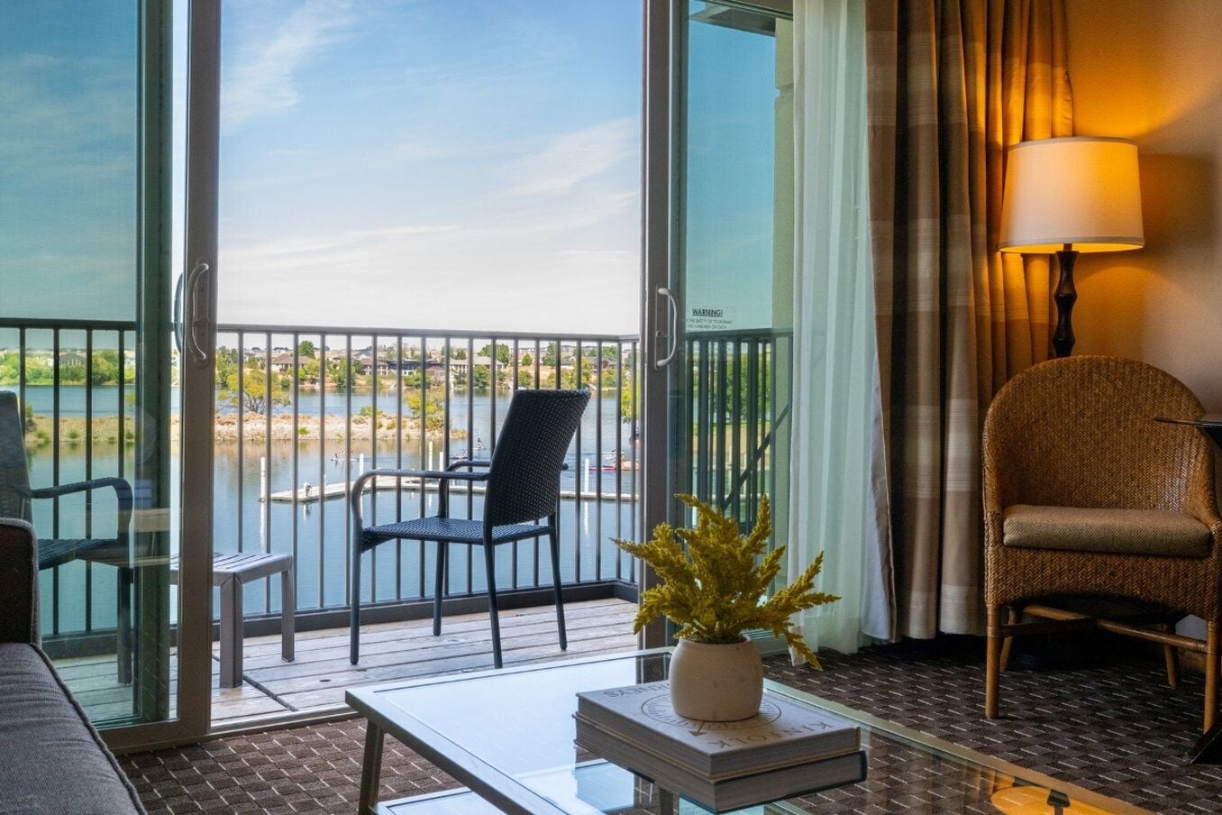 Un balcón con vista a un lago, una silla, una mesa con una planta y una lámpara.