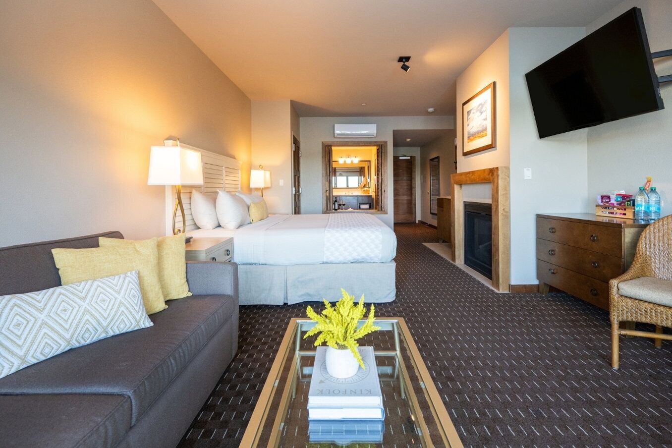 A hotel room with a bed, a sofa, a coffee table with books and a plant, a TV, and a fireplace.